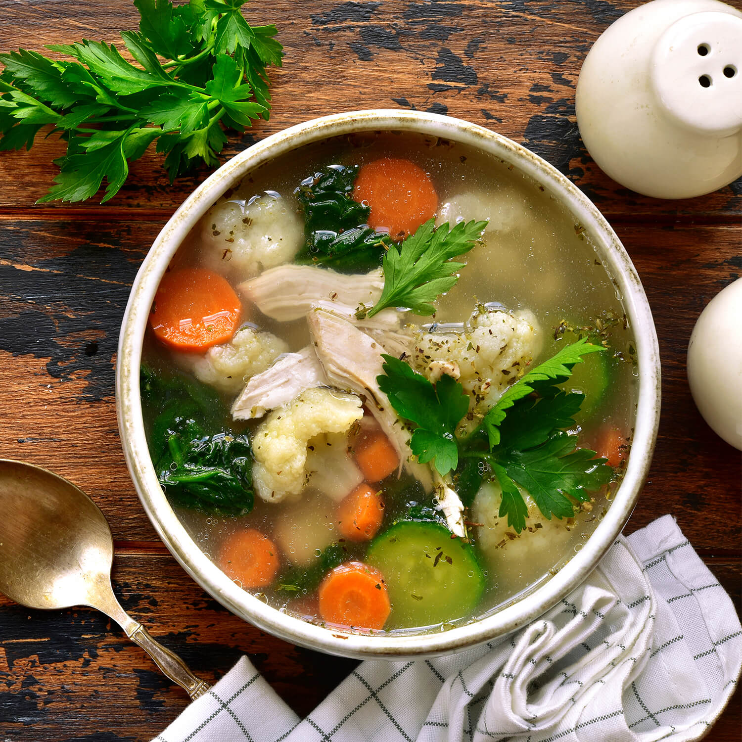 Eine heiße Schüssel Hühnersuppe mit Karotten, Blumenkohl, Spinat und frischer Petersilie, serviert auf einem rustikalen Holztisch