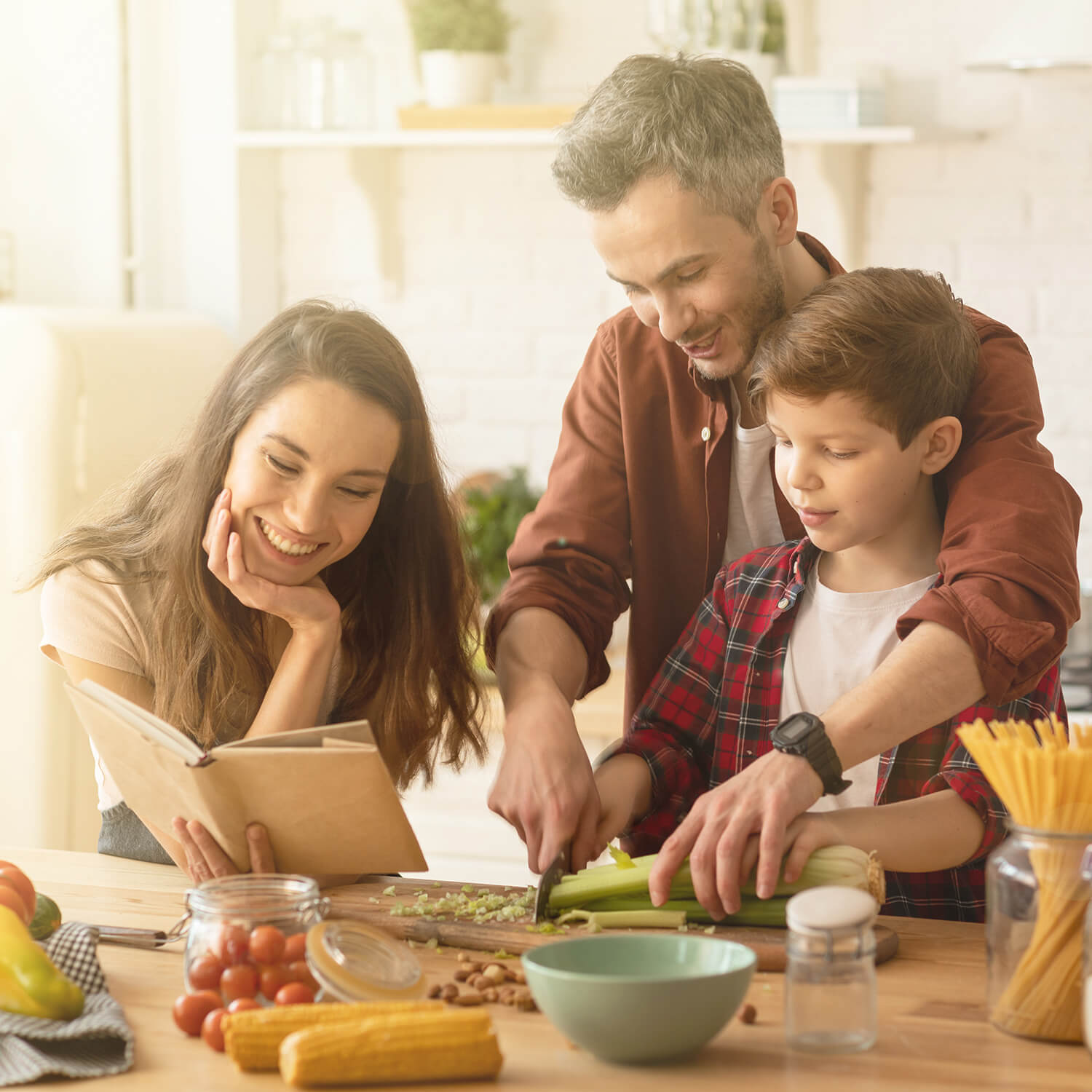 Ein Vater, eine Mutter und ihr Sohn kochen gemeinsam in einer modernen Küche. Der Vater hilft dem Sohn, Gemüse zu schneiden, während die Mutter ein Kochbuch liest.