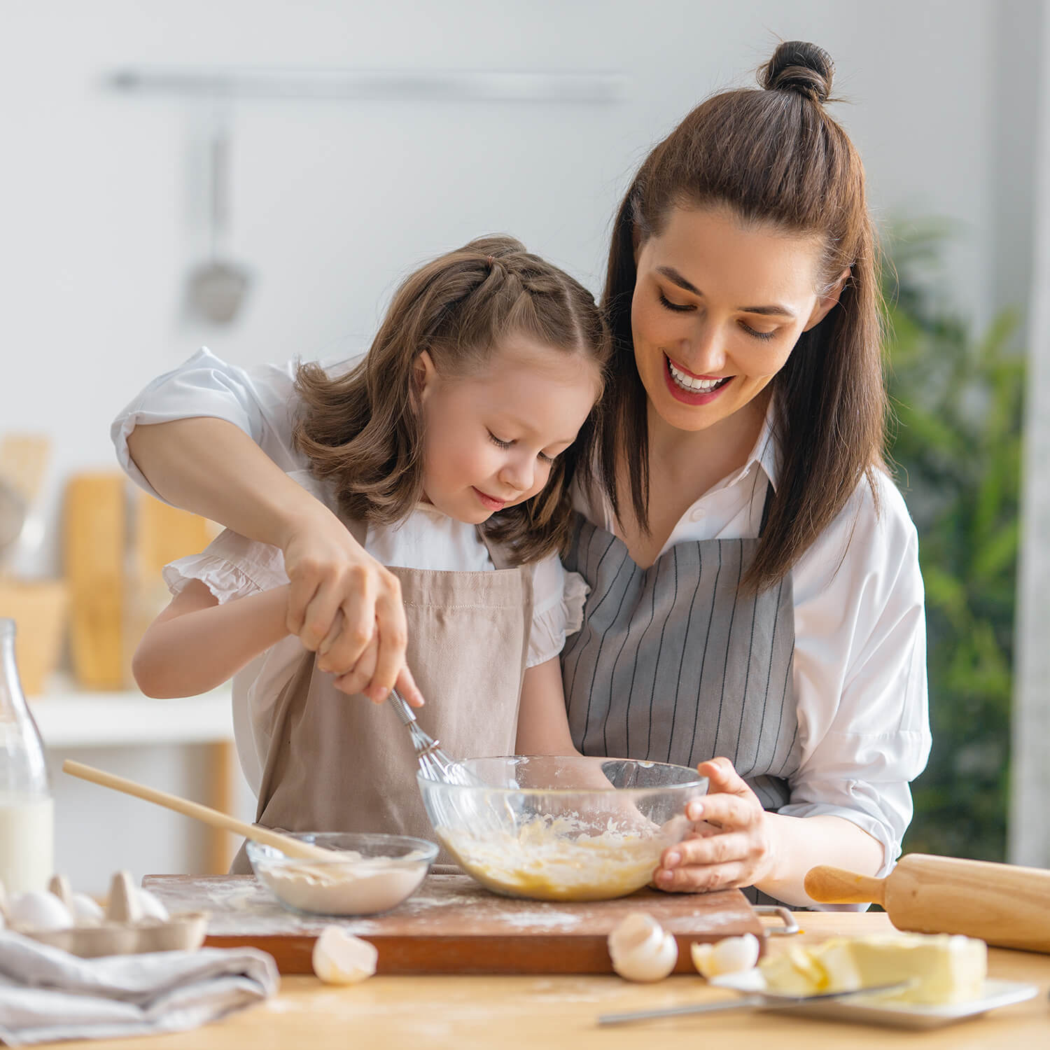 Eine Mutter und ihre kleine Tochter backen gemeinsam in einer hellen, gemütlichen Küche. Beide tragen Schürzen und haben Freude am Rühren des Teigs.